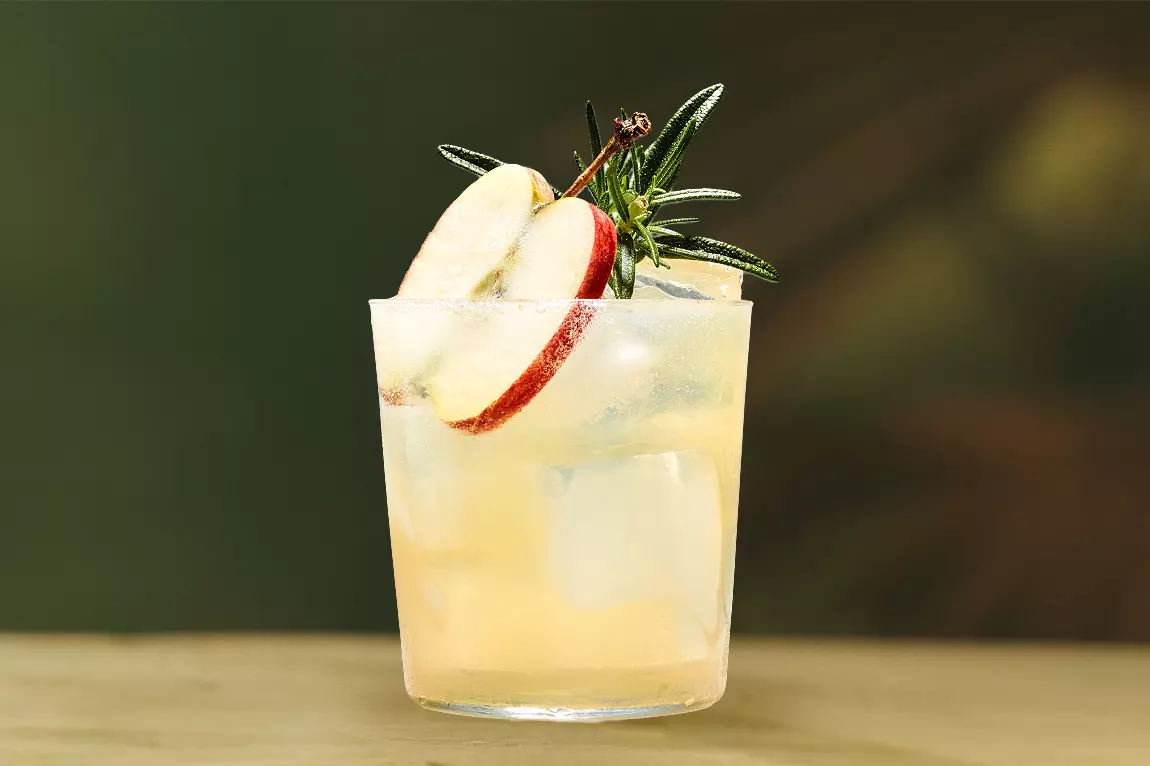 A glass of light-colored iced beverage garnished with apple slices, rosemary, and a clove, set against a blurred green background.