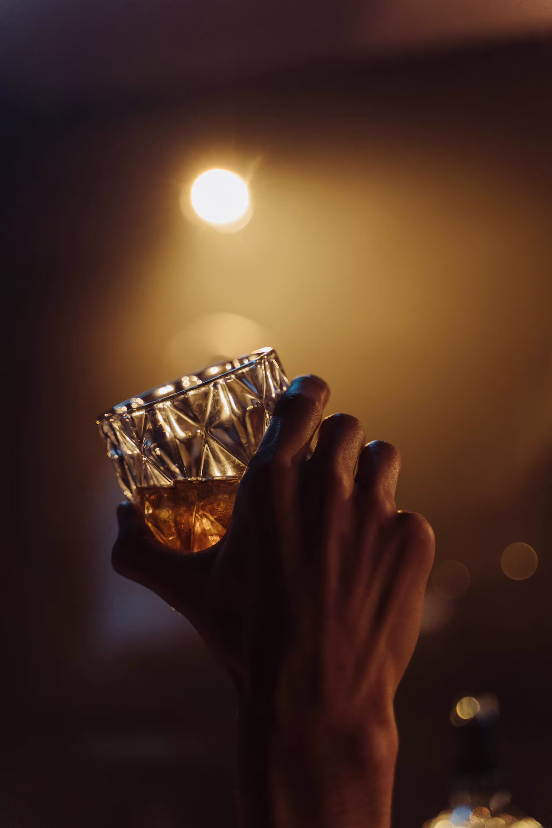A hand holds up a faceted glass of amber liquid, possibly whiskey, against a warm, dimly lit background with a bright light overhead.