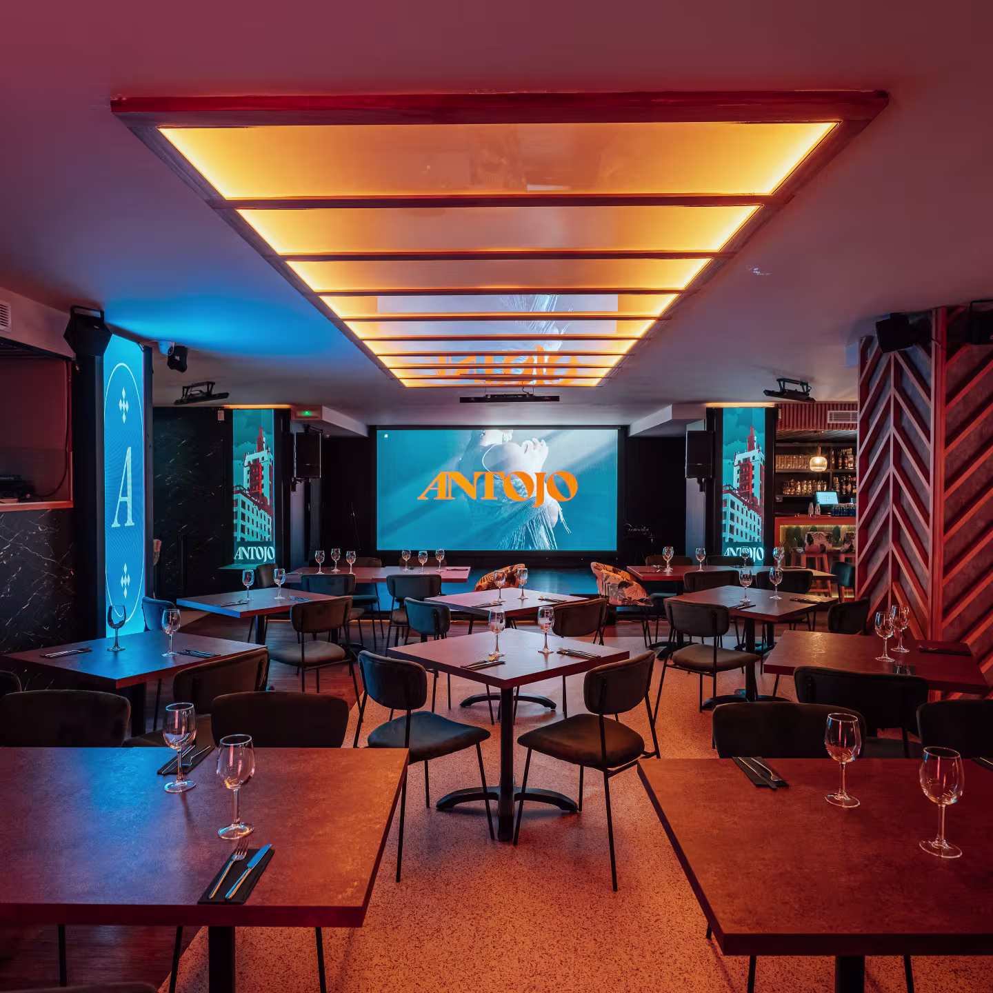 Antojo, espacio moderno con mesas minimalistas, copas listas, iluminación cálida y una pantalla central mostrando su logotipo.