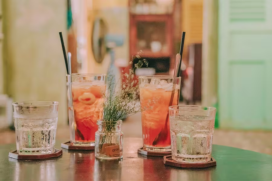 Four glasses with iced drinks and a small glass jar with flowers are arranged on a round table indoors, with a blurred background.