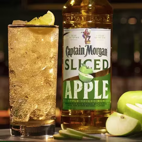 A bottle of Captain Morgan Sliced Apple rum stands next to a tall glass of iced cocktail with a lime wedge, with green apple slices in the foreground.