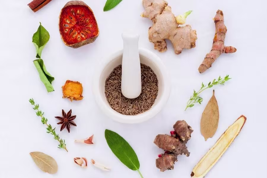A white mortar and pestle with cumin seeds is surrounded by various herbs and spices on a white background.