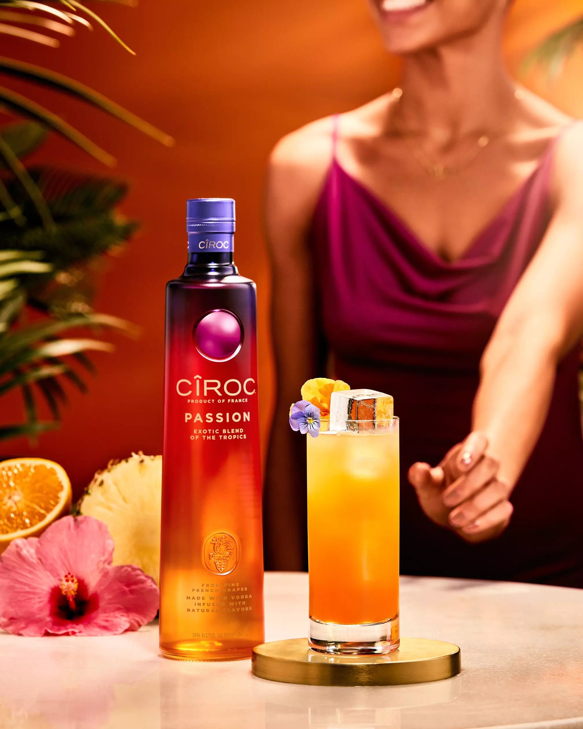 A Cîroc Passion vodka bottle and a colorful cocktail with garnish are placed on a table; a woman in a purple dress is smiling in the background.