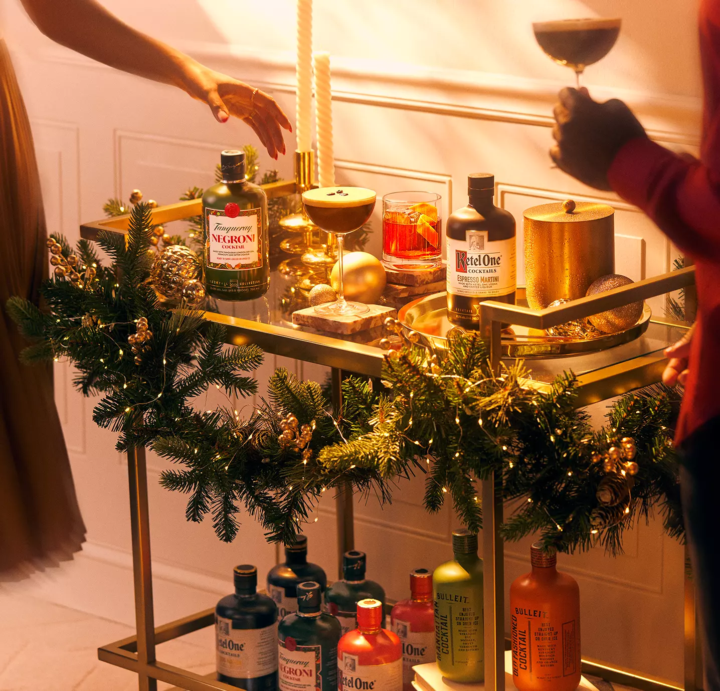 A gold bar cart decorated with holiday greenery holds various bottles of alcohol, glasses, and candles as two people reach for drinks.
