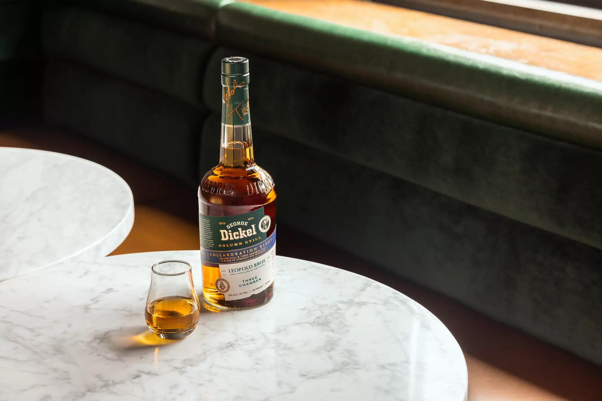 A bottle of George Dickel Tennessee Whisky and a glass with whisky on a round white marble table near a green upholstered bench.