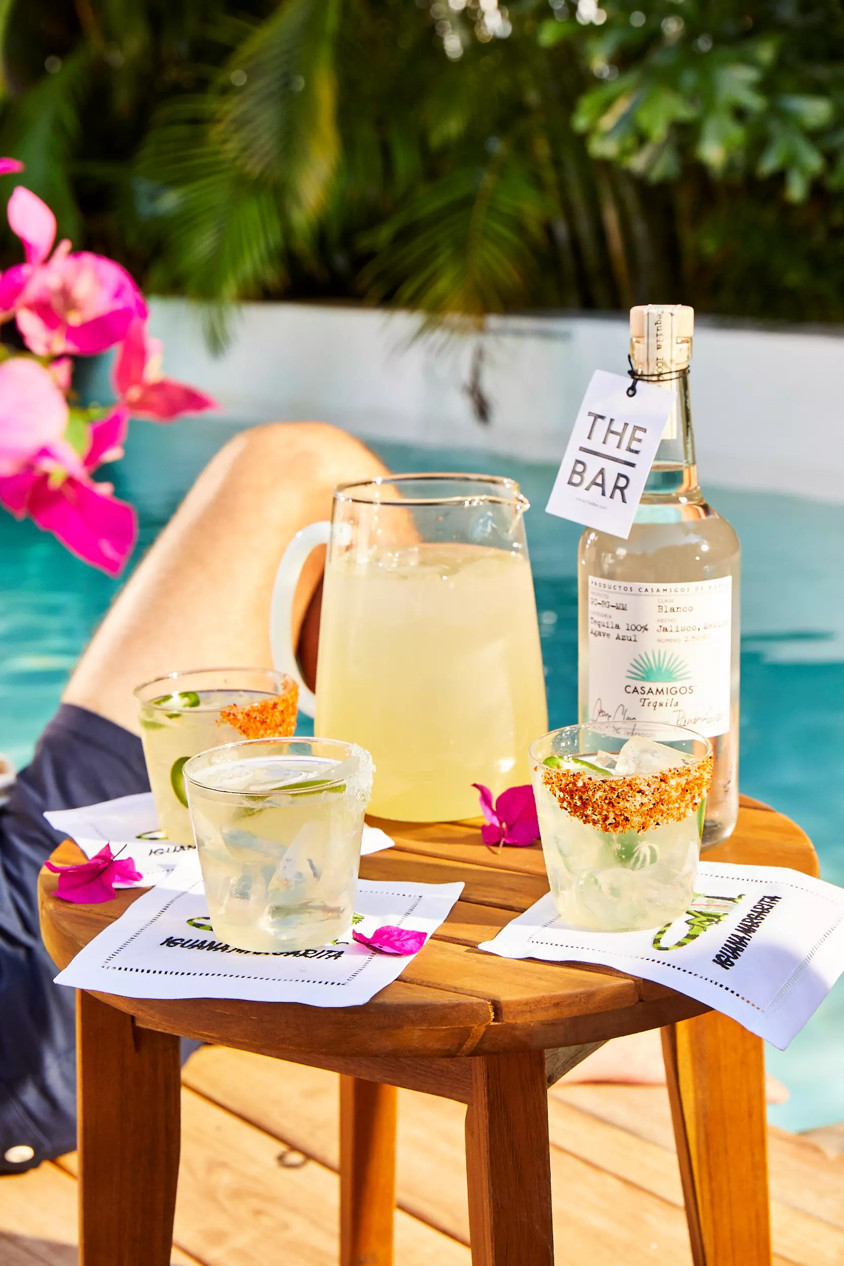 A wooden table by a pool holds a bottle of Casamigos tequila, a pitcher of margarita, three garnished cocktail glasses, and napkins, with a persons legs in the background.