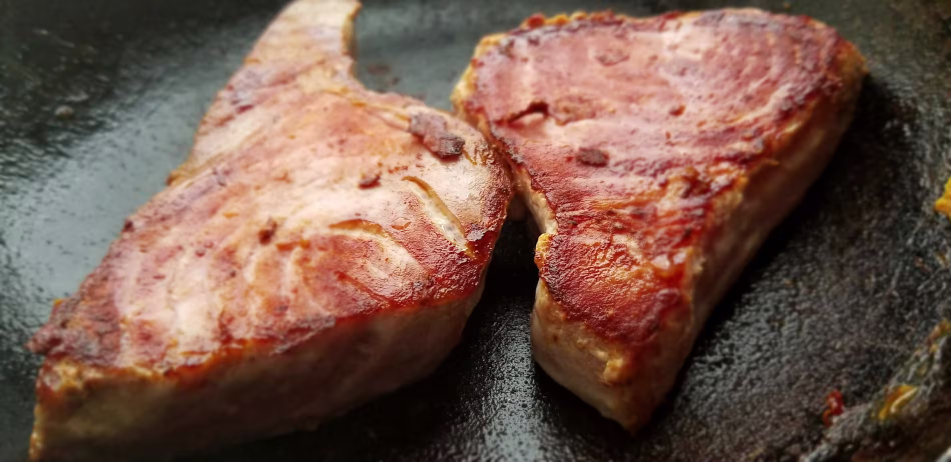 Two seared tuna steaks cooking in a black skillet, showing a browned exterior and visible grill marks.