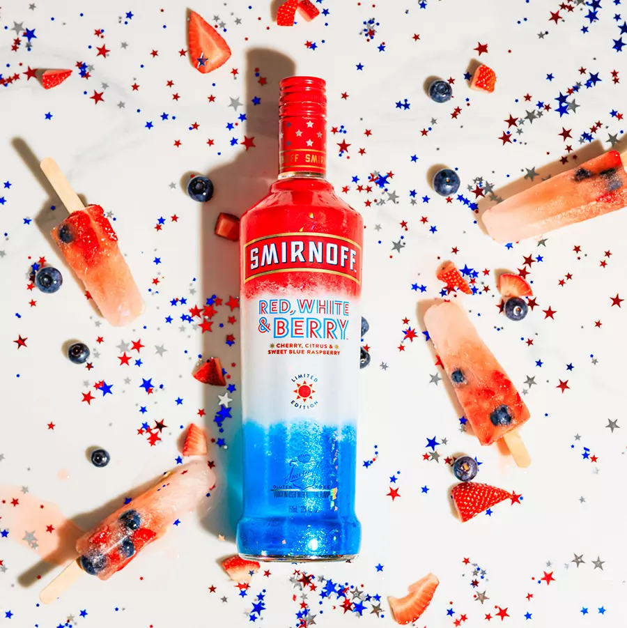 A bottle of Smirnoff Red, White & Berry vodka is surrounded by berry popsicles, strawberries, blueberries, and red, white, and blue confetti stars on a white surface.