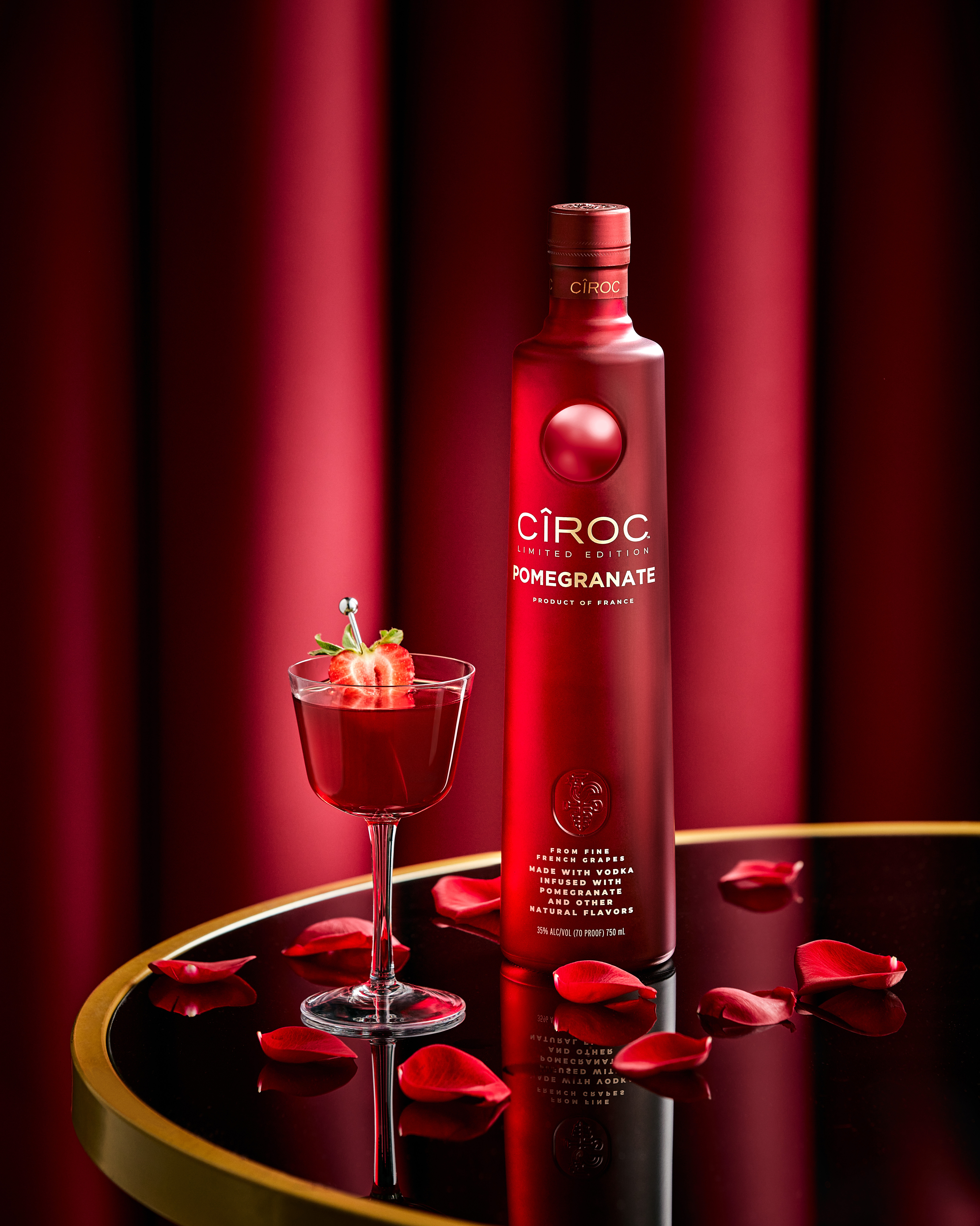 A bottle of Cîroc Pomegranate vodka stands next to a red cocktail garnished with a strawberry, with rose petals scattered on a reflective black and gold table.