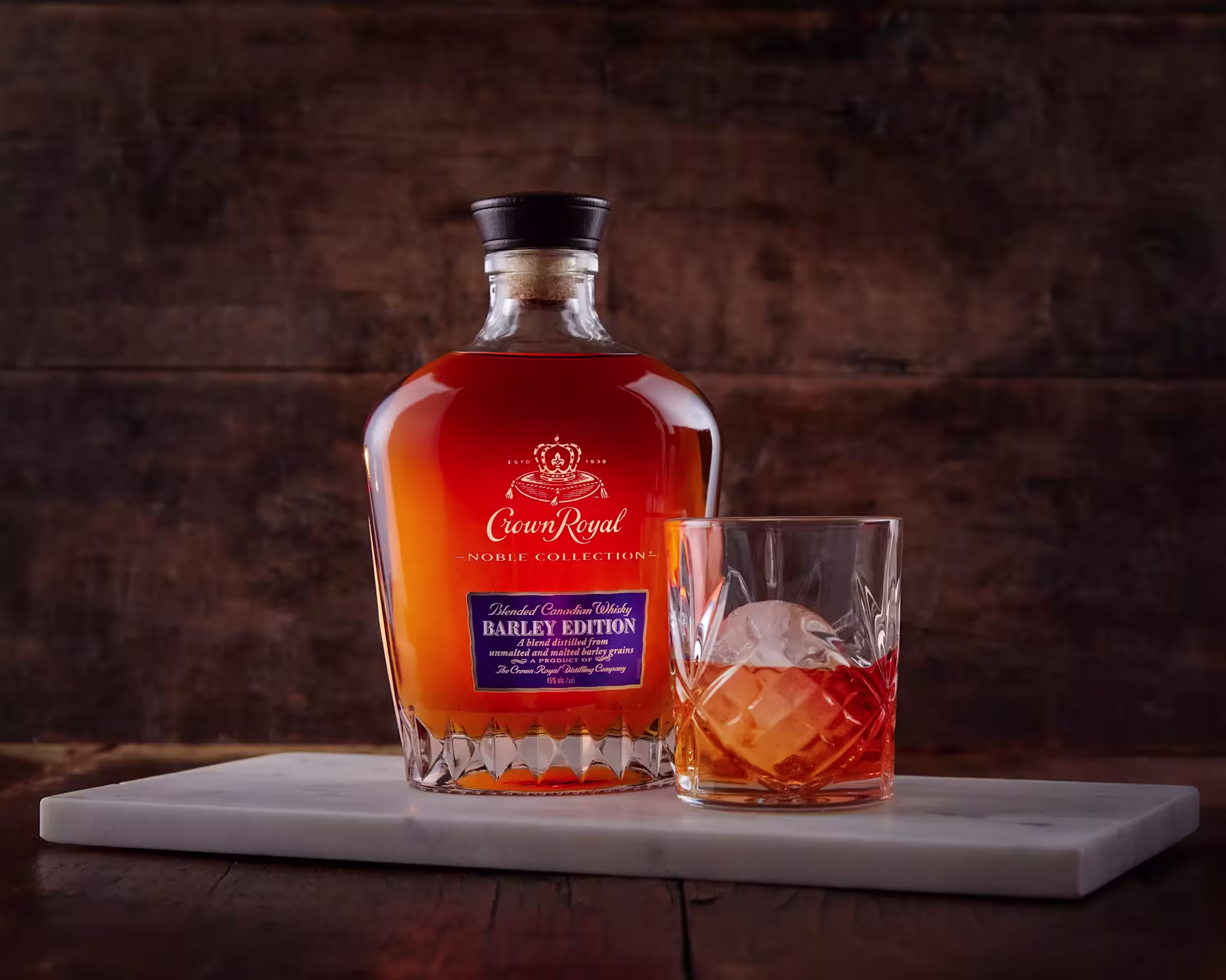 A bottle of Crown Royal Noble Collection Barley Edition whisky sits beside a glass of whisky with a large ice cube on a white marble slab against a wooden background.