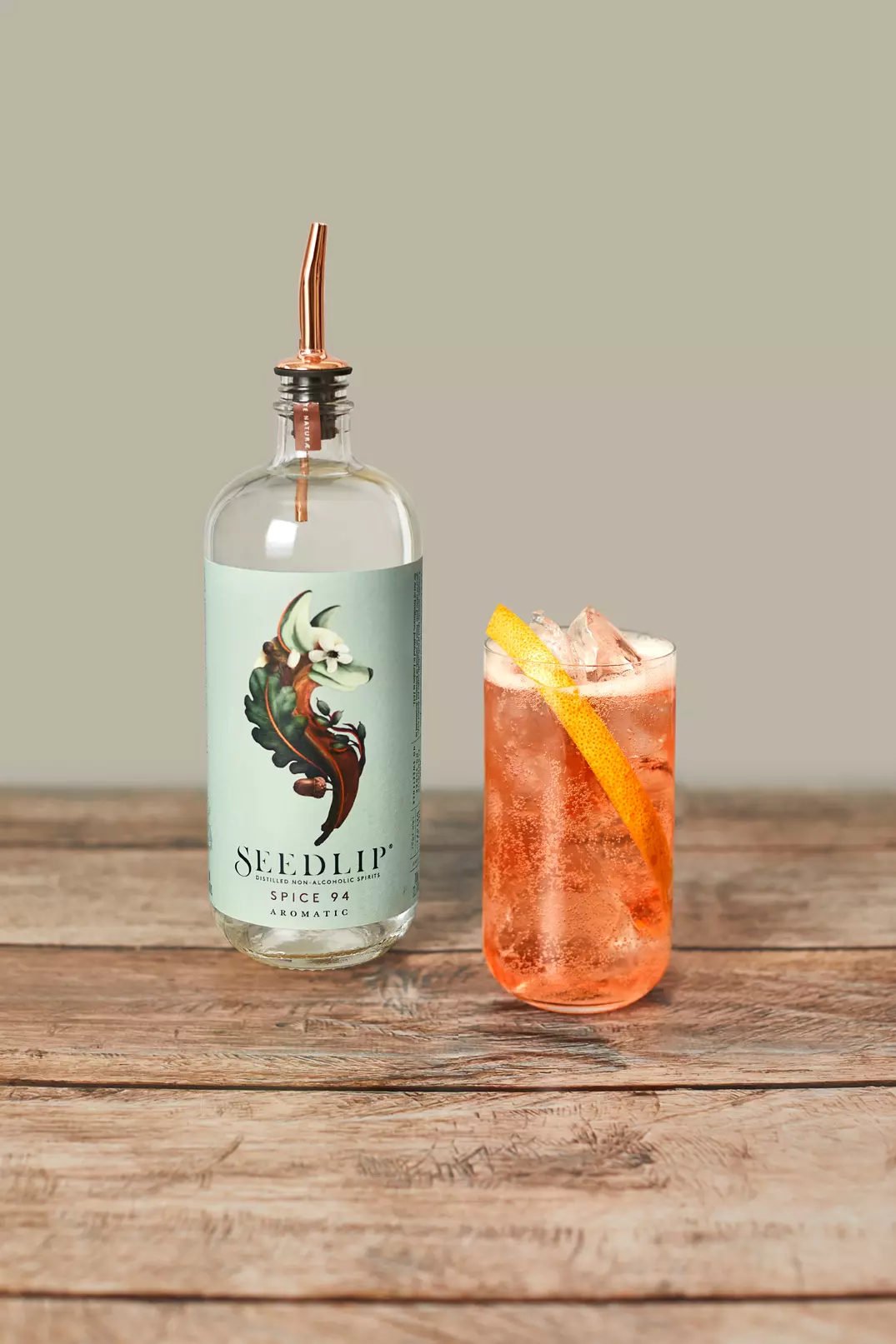 A bottle of Seedlip Spice 94 with a copper pour spout sits next to a glass of iced pink cocktail garnished with a grapefruit slice on a wooden surface.