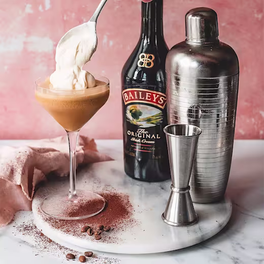 Baileys original bottle beside a cocktail shaker, with a glass of baileys cocktail, being topped with cream