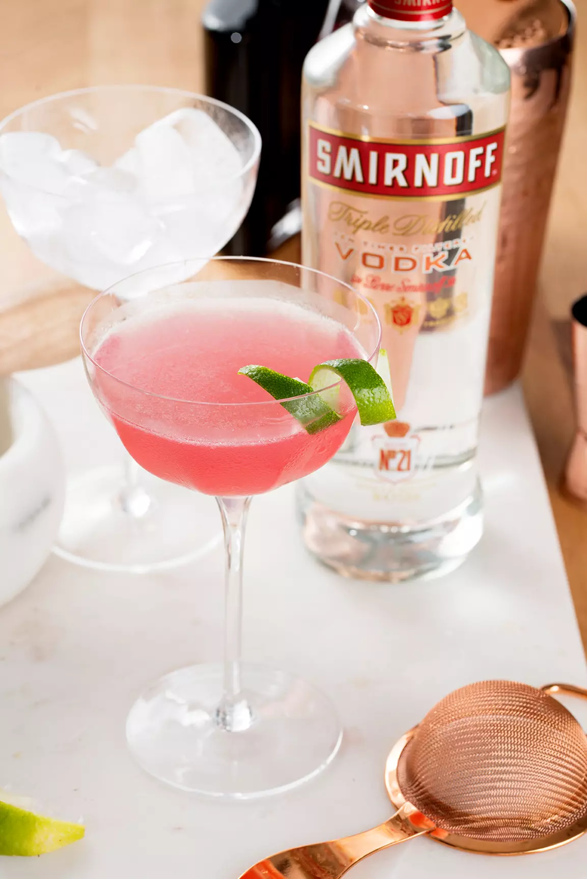 A pink cocktail garnished with a lime twist in a coupe glass, with a Smirnoff vodka bottle, ice-filled glass, and bar tools in the background.