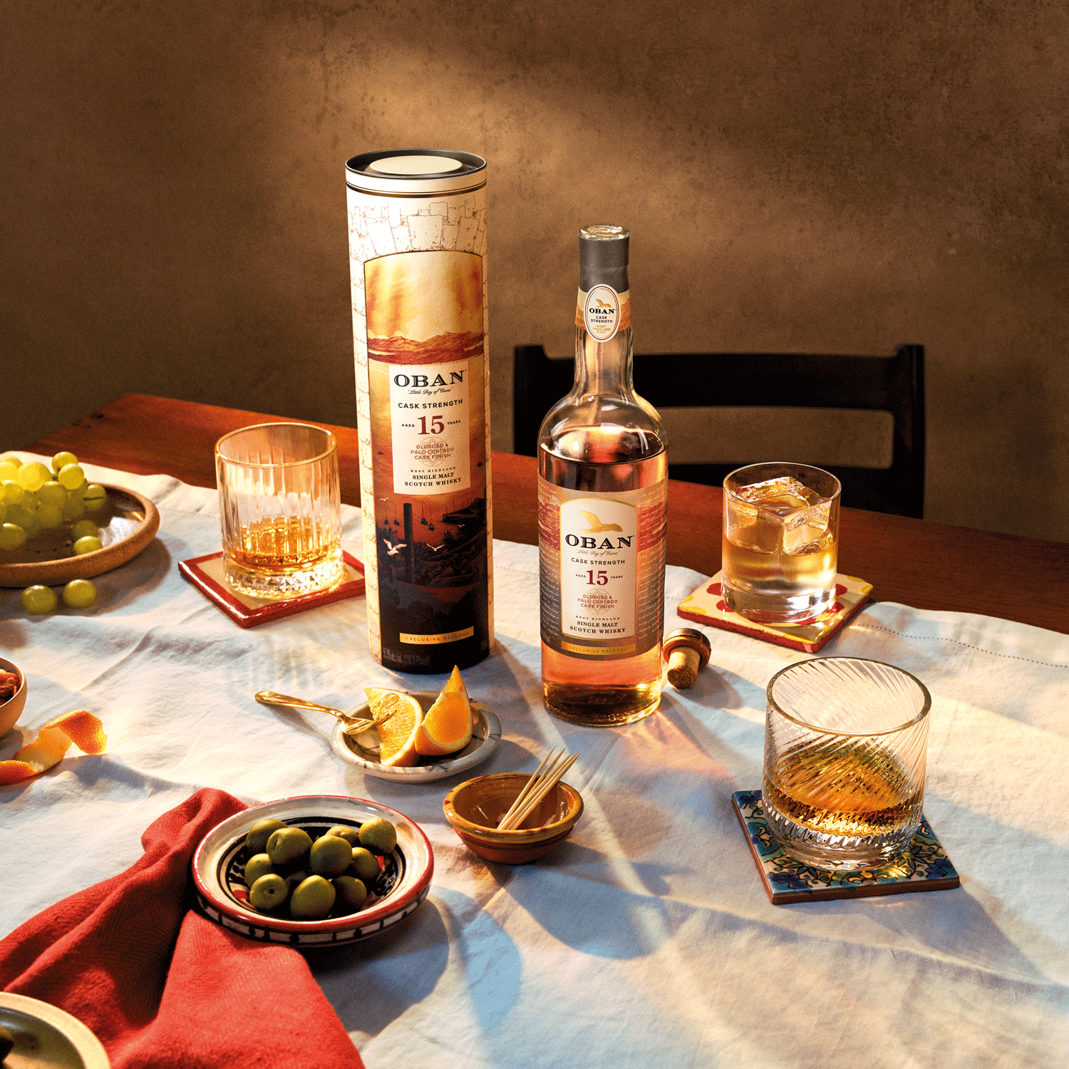 A bottle of Oban 15-year-old whisky with its cylindrical box is on a table set with glasses, olives, orange slices, grapes, and a white tablecloth in warm lighting.