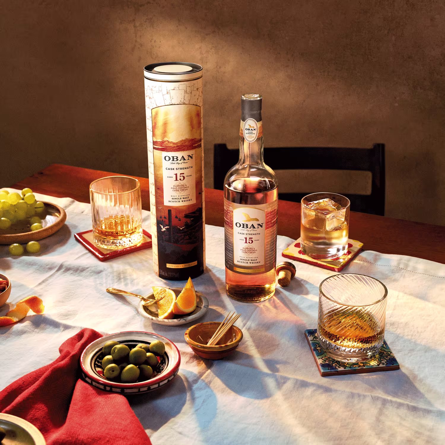 A bottle of Oban 15-year-old whisky with its cylindrical box is on a table set with glasses, olives, orange slices, grapes, and a white tablecloth in warm lighting.