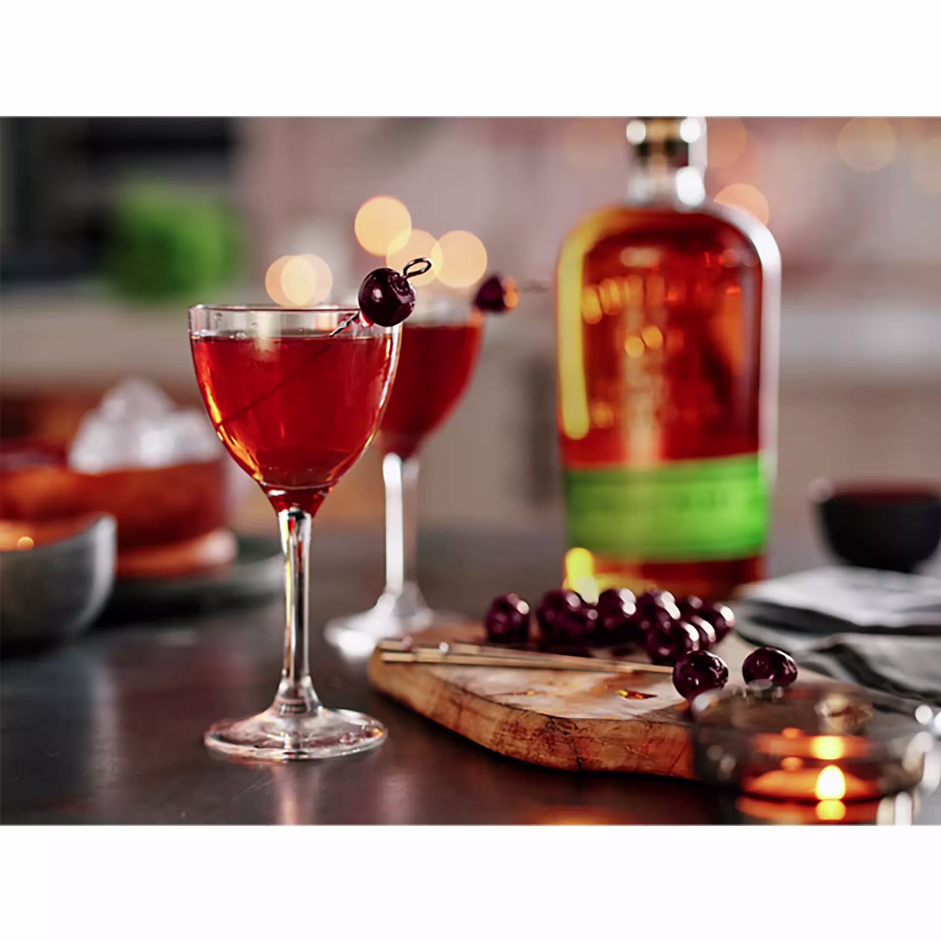 A glass of cocktail garnished with cherries sits on a table next to a bottle of whiskey, a wooden board with cherries, a candle, and blurred background lights.
