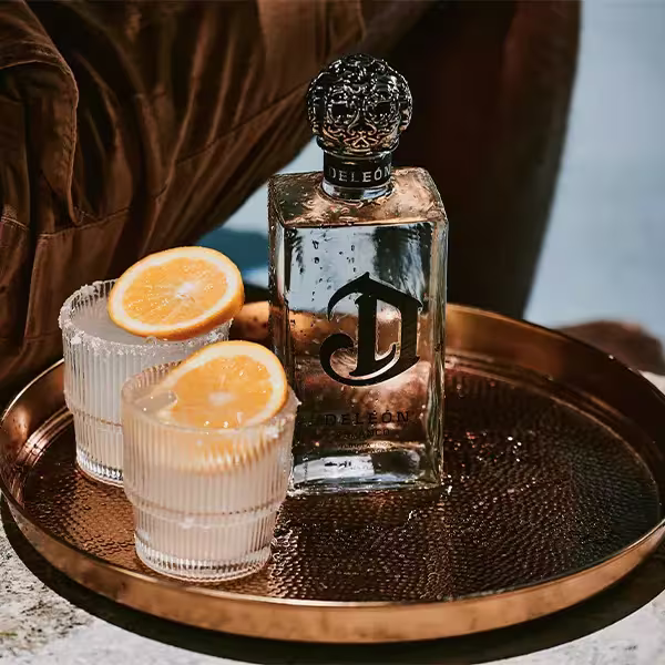 A bottle of DeLeón Tequila and two glasses with ice and orange slices sit on a round brass tray.