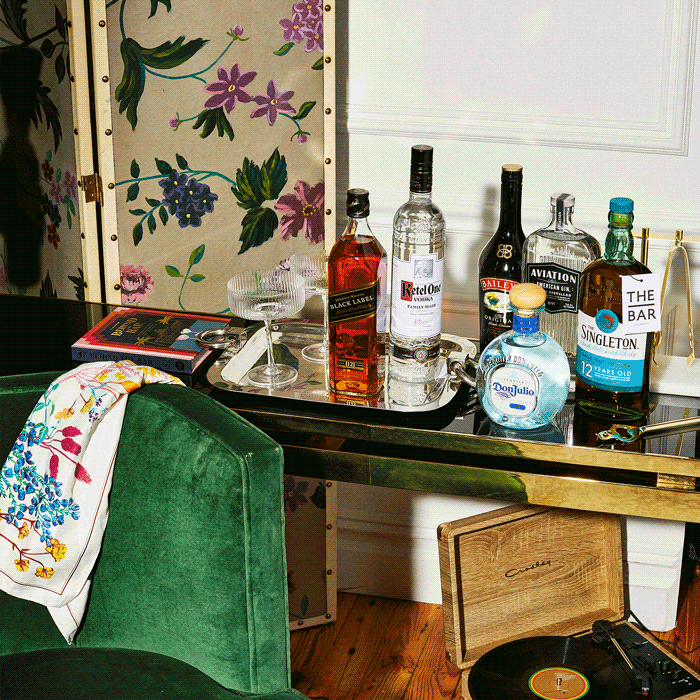 A glass tray on a black and gold table holds bottles of liquor, two glasses, and a book. A green chair, floral screen, and record player are also visible in the scene.