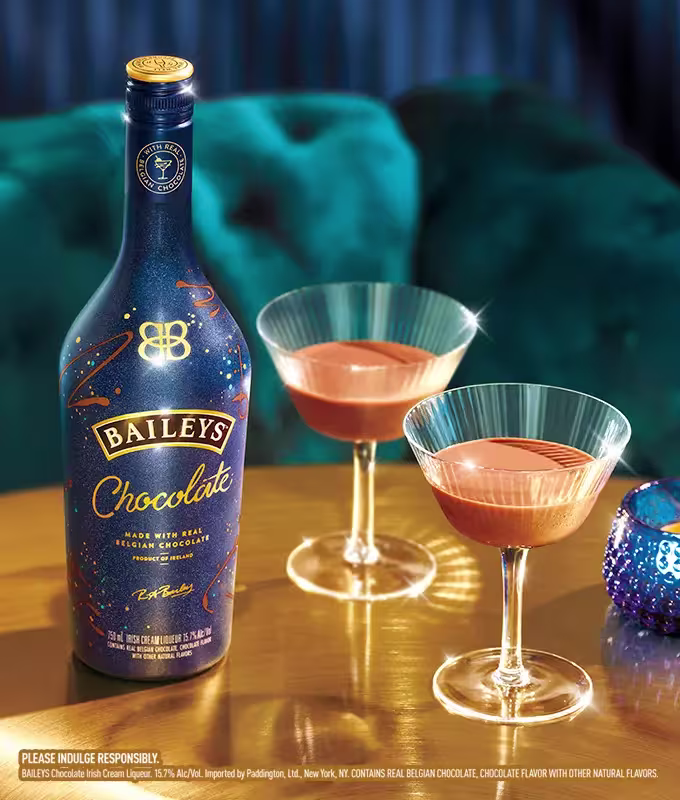 A bottle of Baileys Chocolate liqueur is placed next to two cocktail glasses filled with the drink on a gold surface, with a blue ornament nearby.