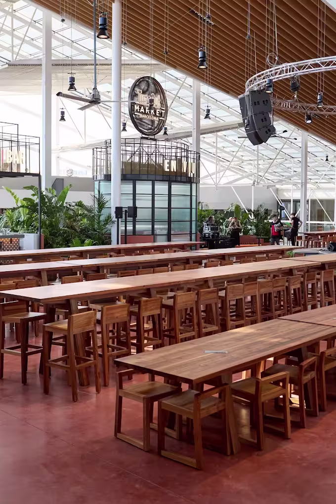 La imagen muestra la terraza de Time Out Market Barcelona, con mesas y sillas modernas, junto a plantas decorativas. Al fondo, se ve un mural con el logo de Time Out Market y una vista hacia la ciudad.