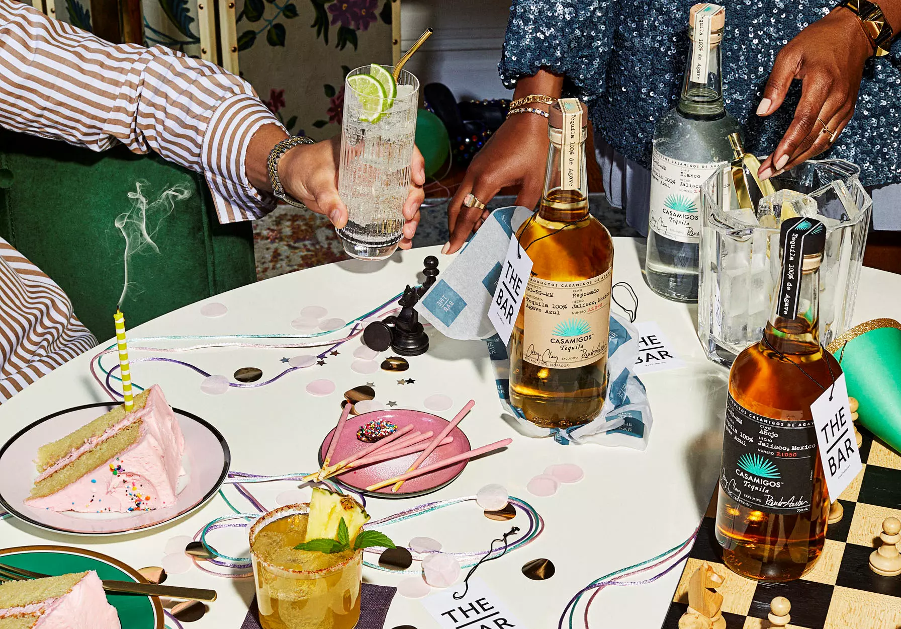 A table with cocktails, bottles of alcohol, a slice of cake, party decorations, and two people clinking glasses.
