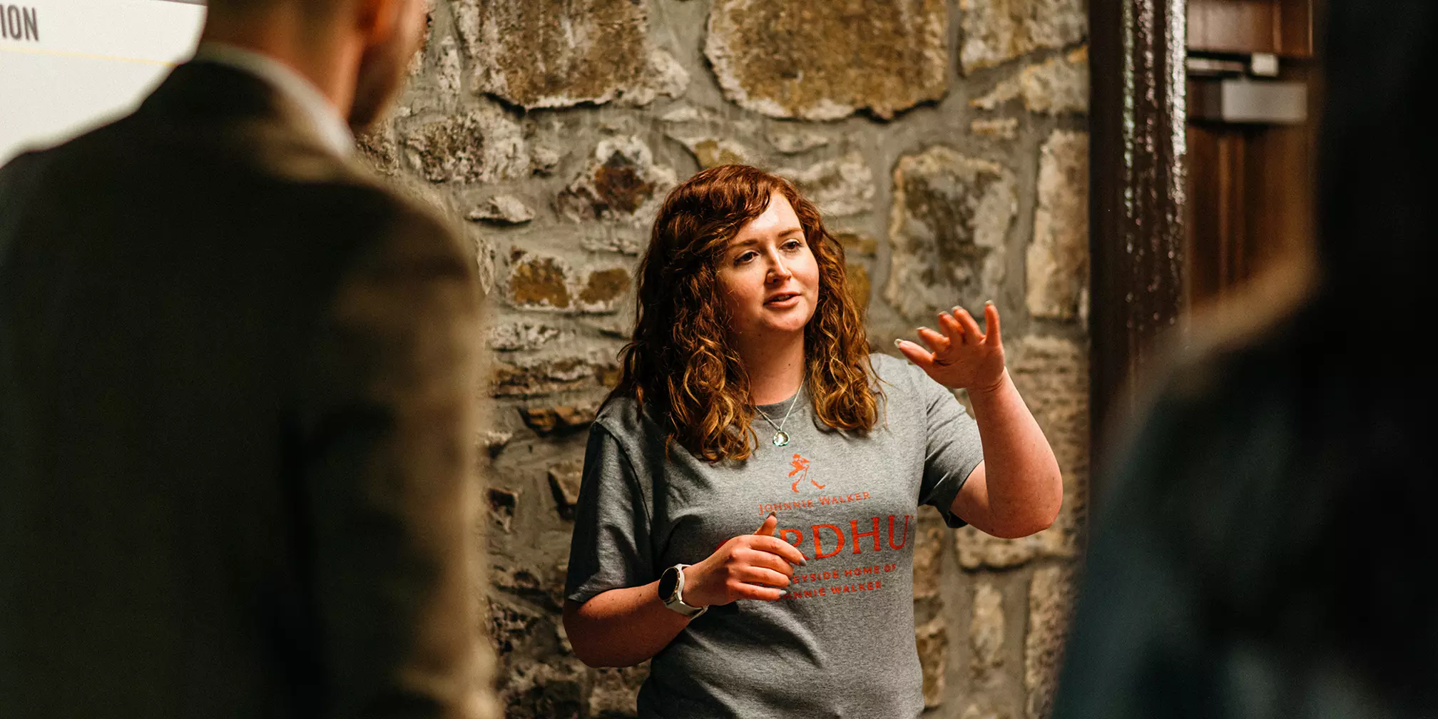 A woman with curly hair speaks and gestures with one hand, standing in front of a stone wall while two people listen.