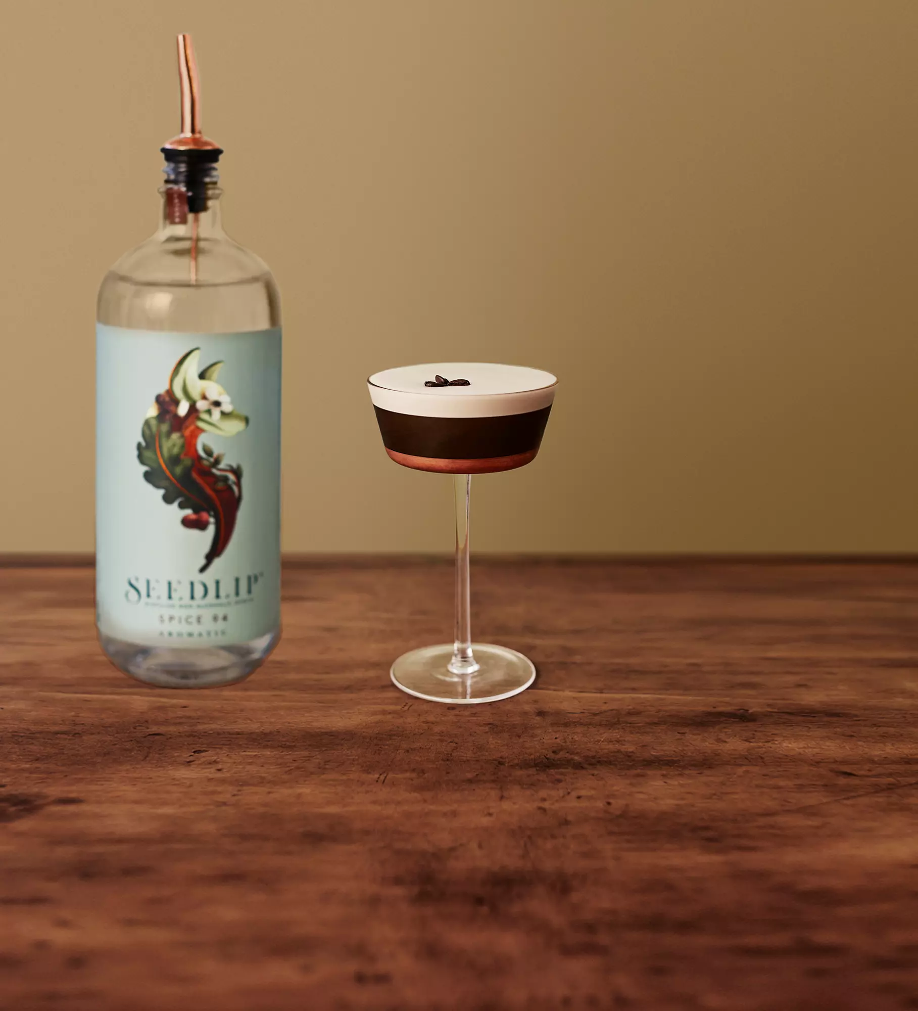 A bottle labeled Seedlip Spice 94 with a copper pour spout next to a coupe glass containing a dark cocktail topped with foam and three coffee beans, on a wooden surface.