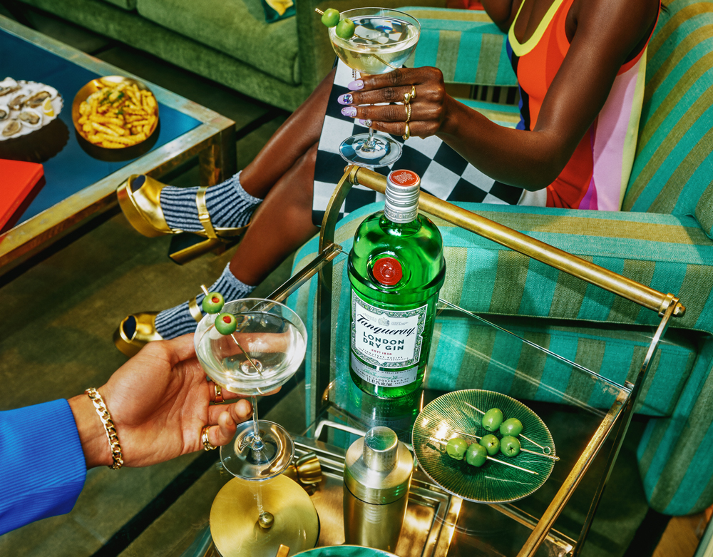 Two people holding martini glasses garnished with olives, seated near a cart with a gin bottle, cocktail shaker, olives, and snacks on nearby tables.