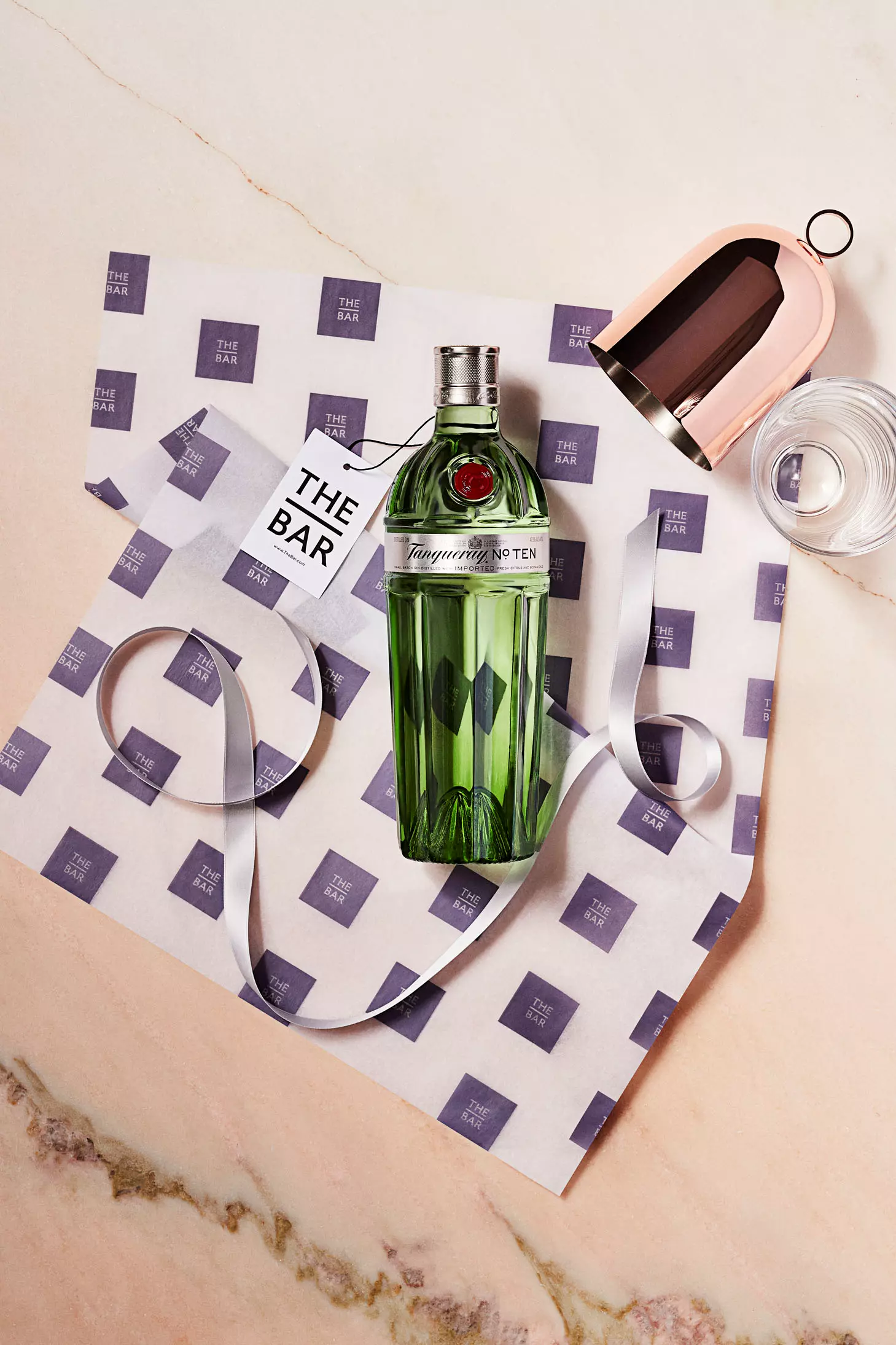 A bottle of Tanqueray No. Ten gin on branded paper with a ribbon, a cocktail shaker lid, a glass of water, and a card reading The Bar on a marble surface.