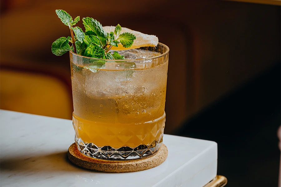 A glass of iced amber-colored cocktail garnished with fresh mint and a slice of dried fruit, placed on a coaster on a marble table.