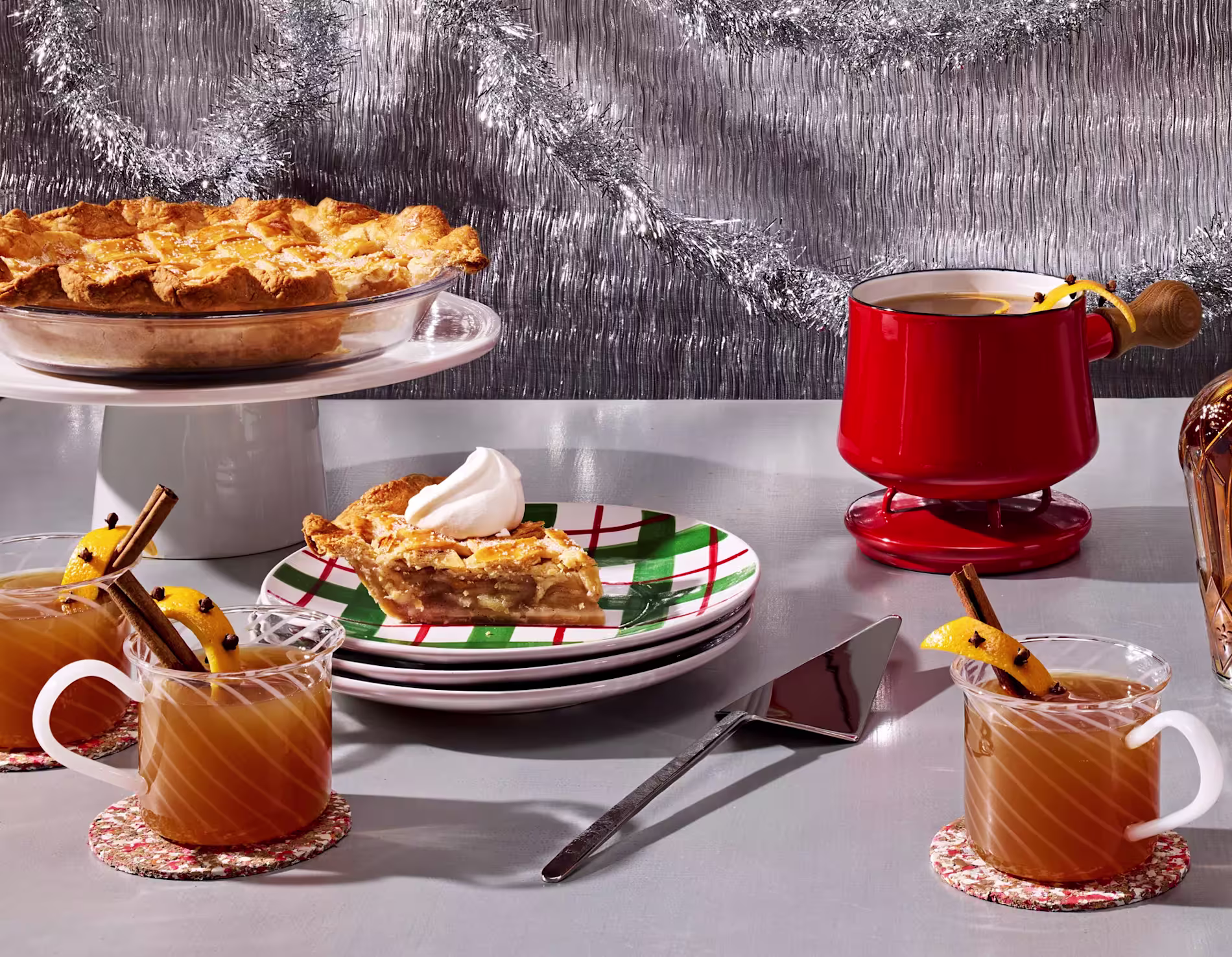 A table with apple pie, a slice served on a plate with whipped cream, three mugs of spiced drink with cinnamon sticks, and a red mug, set against a silver tinsel backdrop.