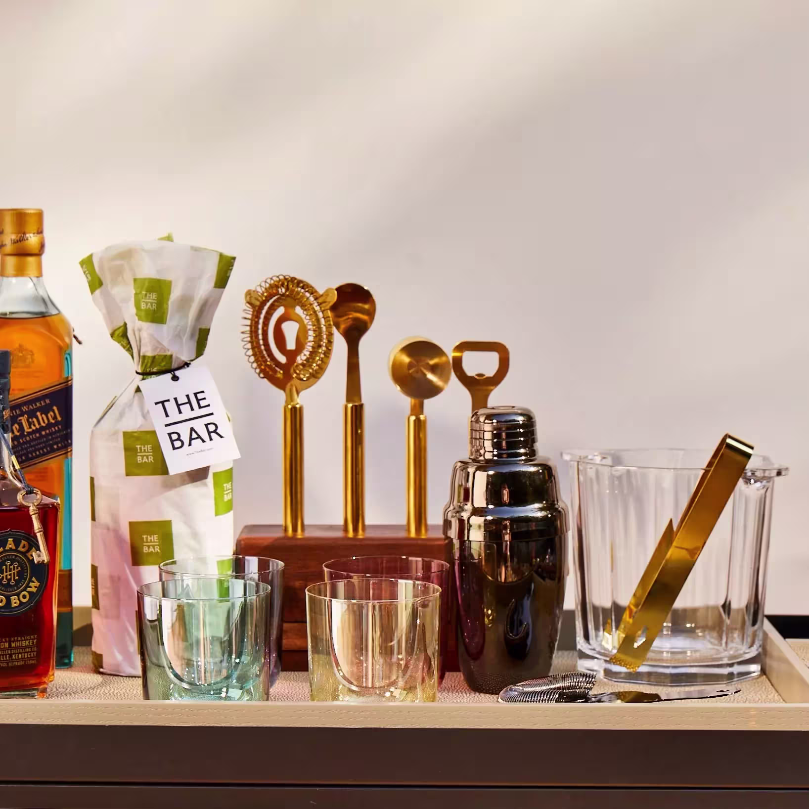 A bar setup with whiskey bottles, wrapped gift, various cocktail tools, shaker, ice bucket with tongs, strainer, and three drinking glasses on a tray.