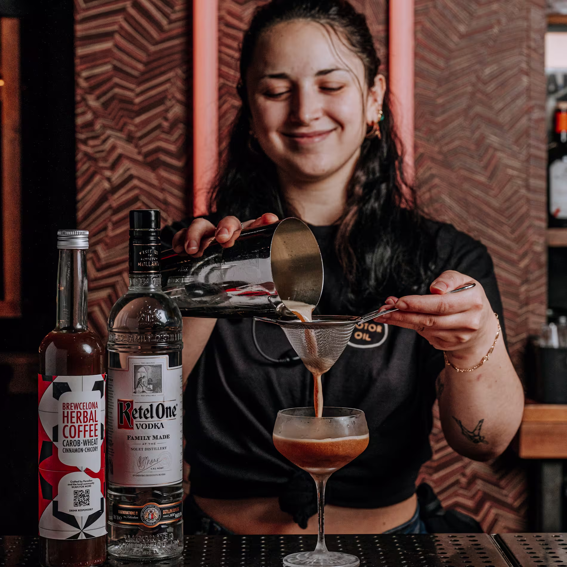 Bartender sonriente filtrando un cóctel en copa sobre la barra. Frente a ella, botellas de Ketel One y Brewcelona Herbal Coffee. Fondo con patrón geométrico y tubos de neón rosa.