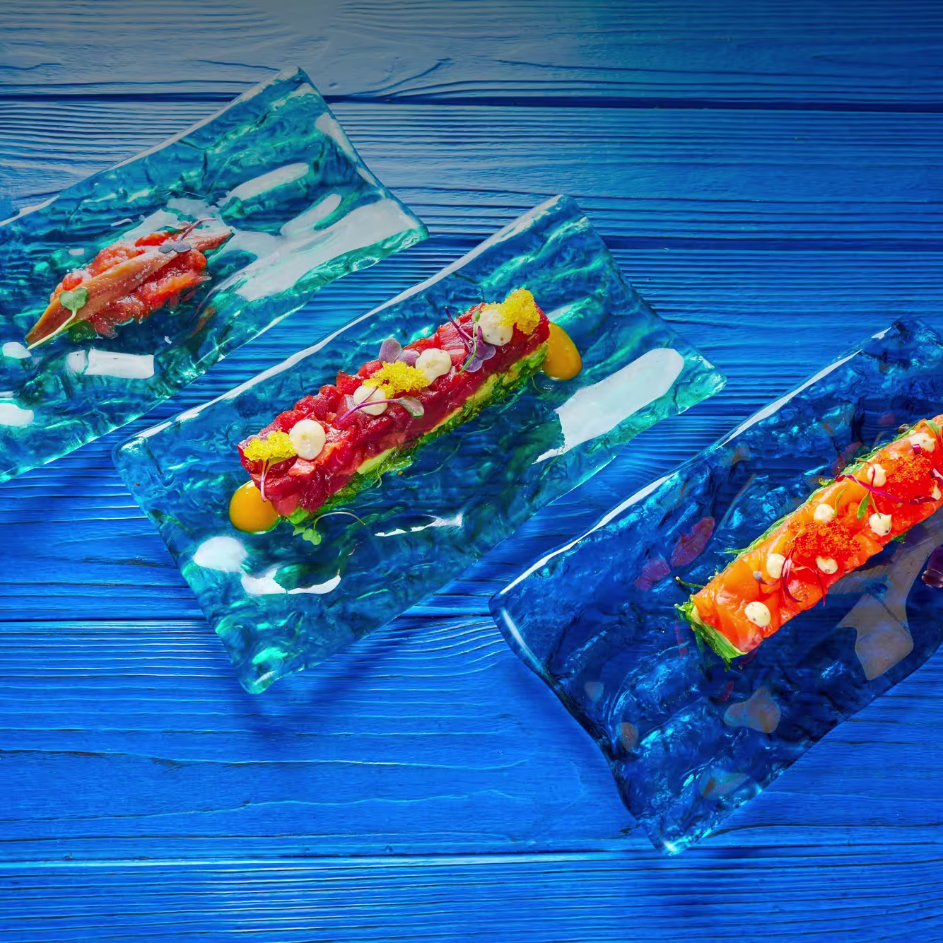 Tres platos de pescado crudo servidos con colorido y precisión sobre bandejas de cristal azul, sobre una mesa que simula el fondo del mar.