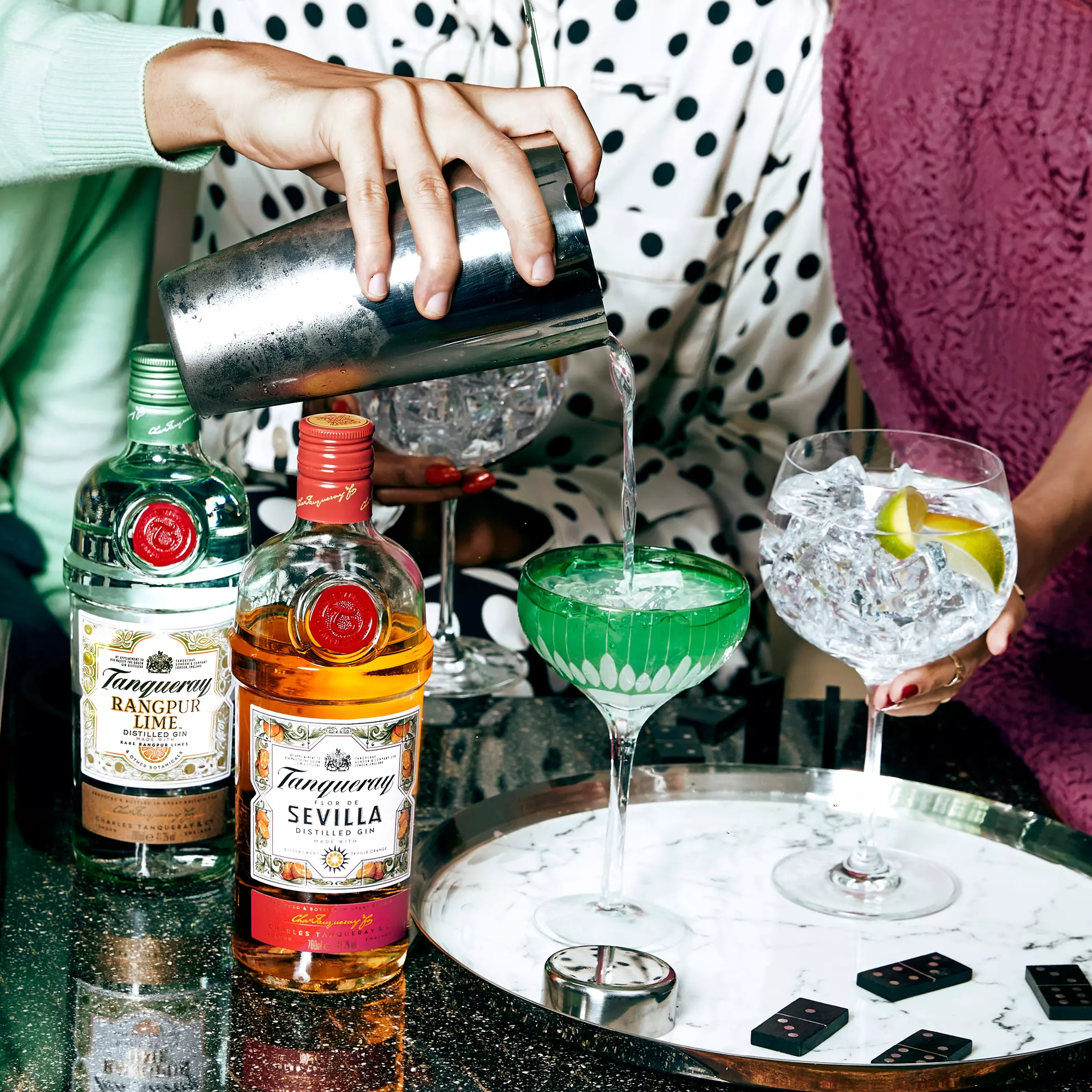 A person pours a green cocktail from a shaker into a coupe glass, with Tanqueray gin bottles, gin and tonic glasses, and playing cards on a table.