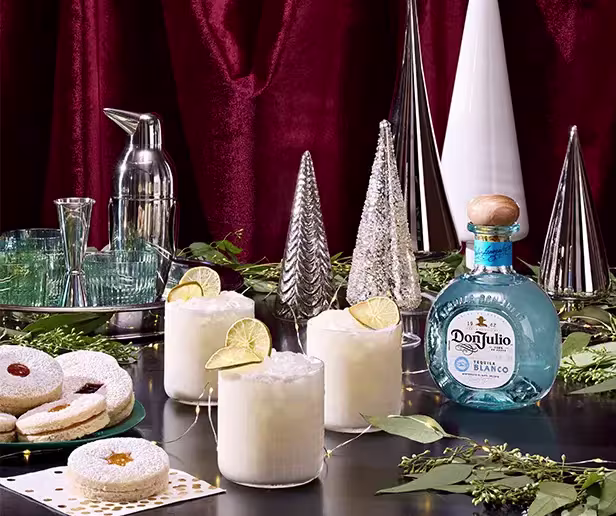 A table with three cocktails garnished with lime, a bottle of Don Julio tequila, cookies, glassware, metallic cone decorations, and greenery against a red curtain backdrop.