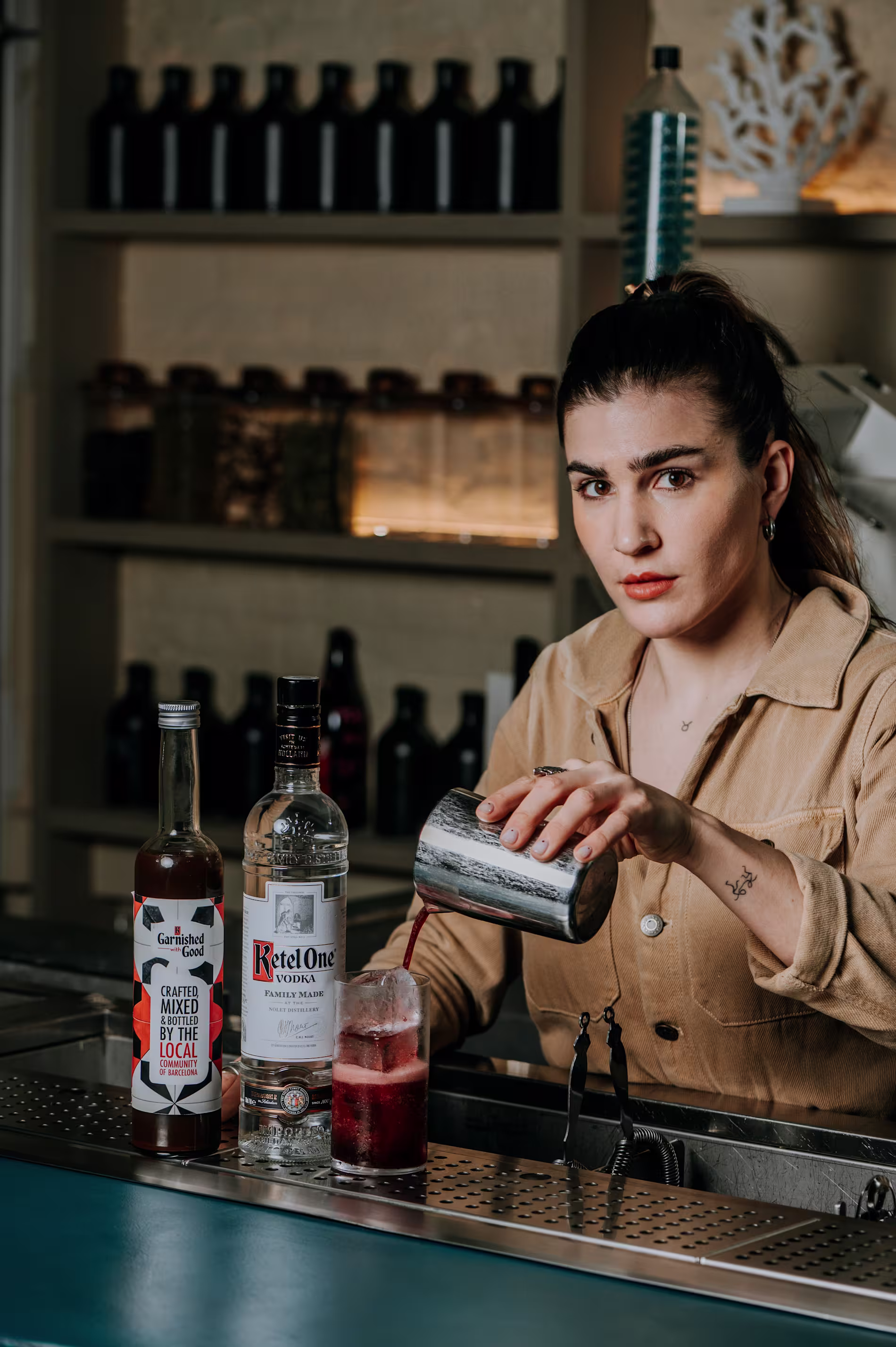 Bartender sirviendo un cóctel rojo en vaso con hielo. Frente a ella, botellas de Ketel One y Garnished with Good. Al fondo, estanterías con botellas oscuras y tarros iluminados.