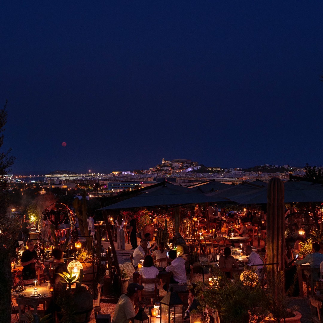 En Casa Maca, disfruta de una atmósfera mágica con vistas nocturnas a Ibiza, rodeado de plantas y mesas iluminadas por velas.