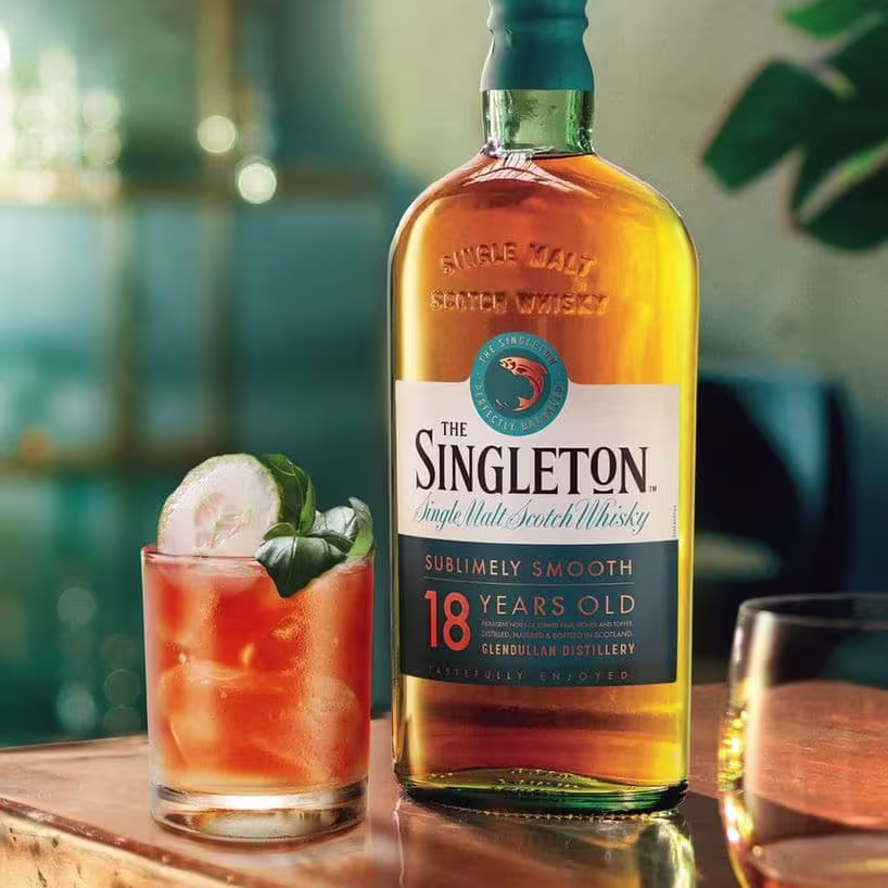 O sticlă de Single Malt Scotch Whisky The Singleton 18-Year-Old stă lângă un cocktail garnisit cu castravete și busuioc. Pe etichetă scrie "Sublimely Smooth". Decorul este o cameră caldă, slab luminată, sugerând o atmosferă primitoare pentru savurarea whisky-ului.