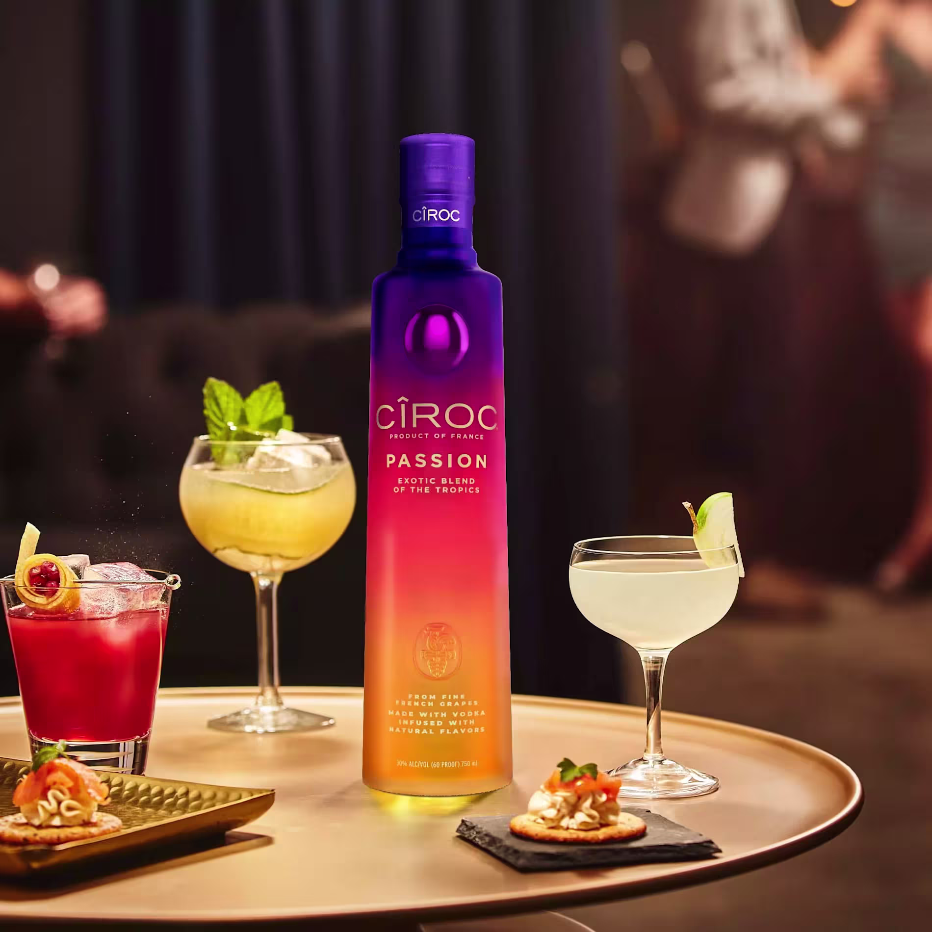 A bottle of Ciroc Passion vodka is displayed on a table with three cocktails and appetizers; blurred people are visible in the background.
