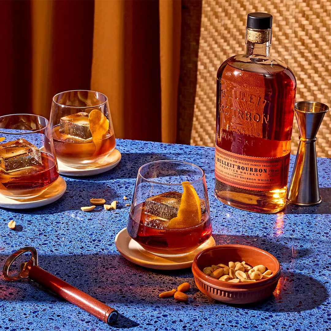 A bottle of Bulleit Bourbon, three whiskey glasses with ice and orange peel, a jigger, a bowl of mixed nuts, and a bottle opener on a blue speckled table.