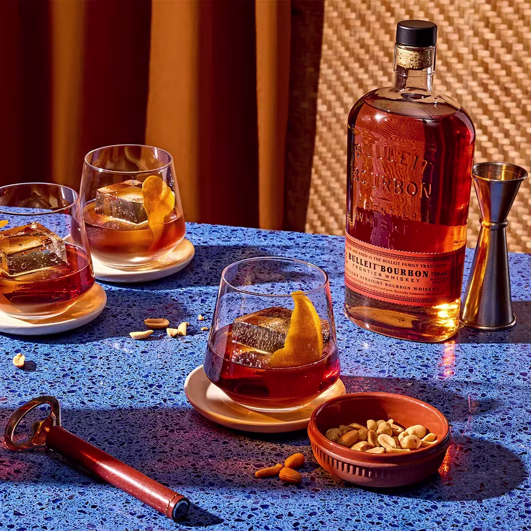 A bottle of Bulleit Bourbon, three whiskey glasses with ice and orange peel, a jigger, a bowl of mixed nuts, and a bottle opener on a blue speckled table.