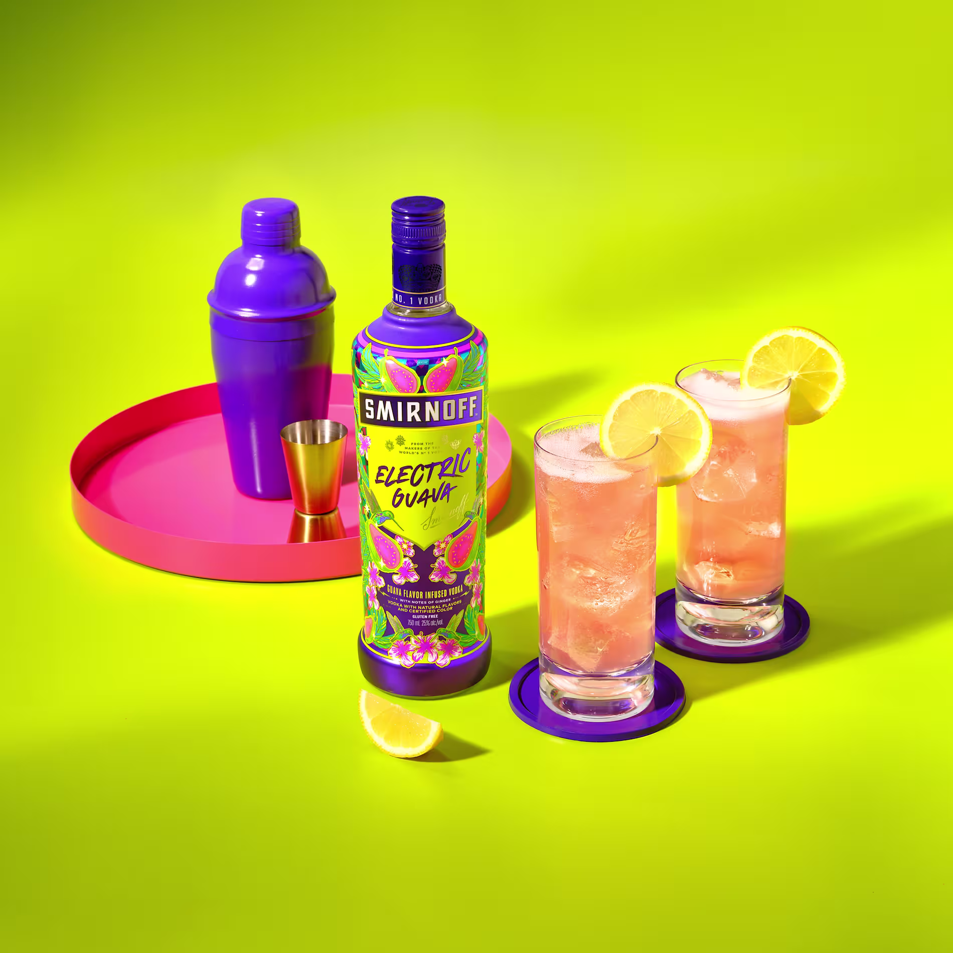 A bottle of Smirnoff Electric Guava, two cocktails with lemon slices, a purple cocktail shaker, a jigger, and a lemon wedge on a pink tray against a bright yellow background.