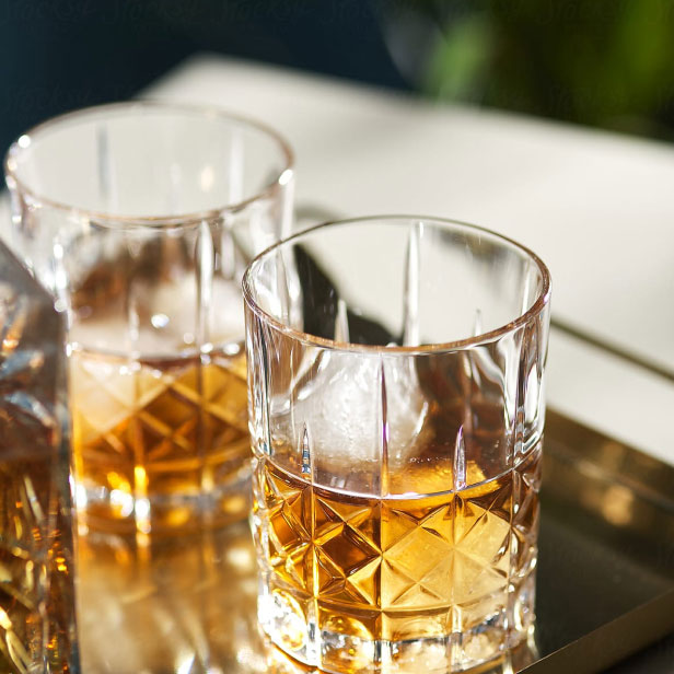 A close-up of a glass tumbler containing a brown liquid, likely whiskey or bourbon, placed on a reflective surface with sunlight shining on it.