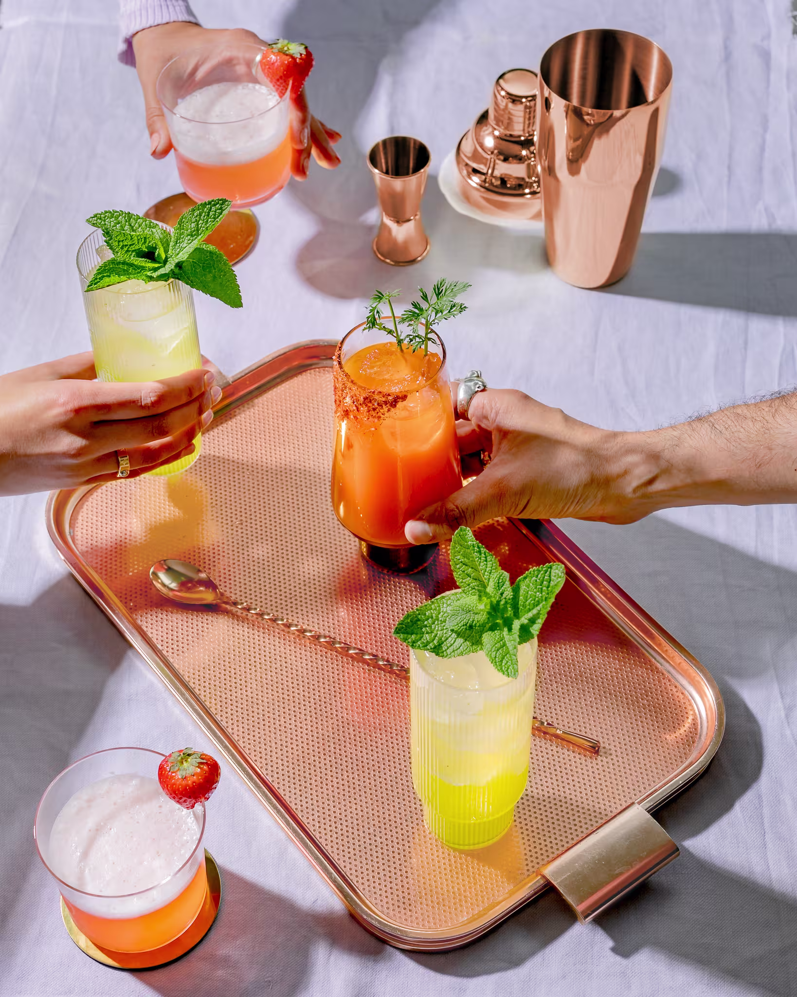 Hands holding vibrant cocktails garnished with fresh herbs, surrounded by a copper tray and cocktail tools.