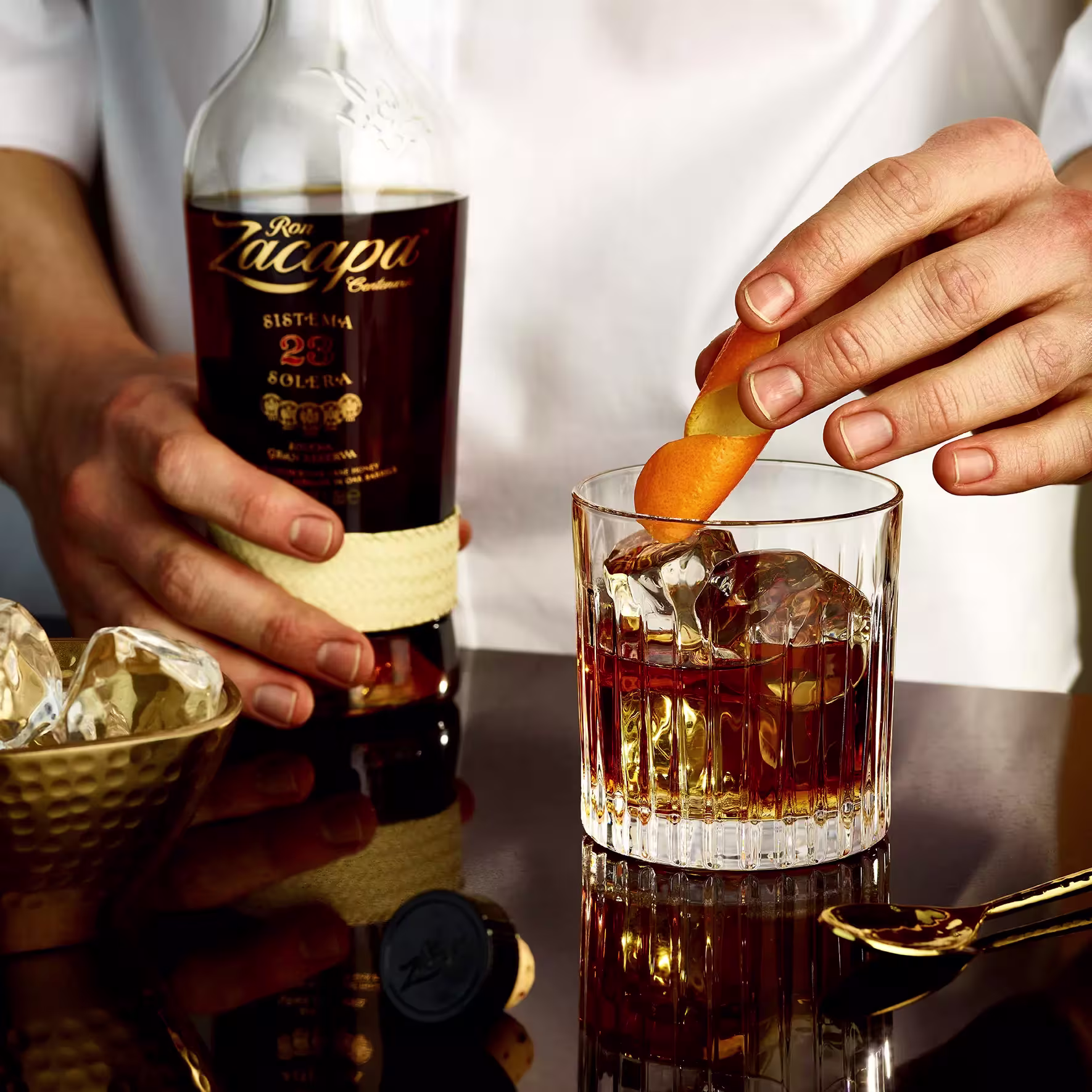 A person garnishes a glass of whiskey with an orange peel while holding a bottle of Ron Zacapa 23 rum; ice and bar tools are nearby on the table.