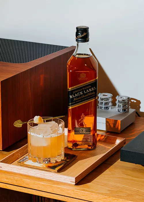 A bottle of Johnnie Walker Black Label whisky and a cocktail glass with ice sit on a wooden tray on a table next to audio equipment.