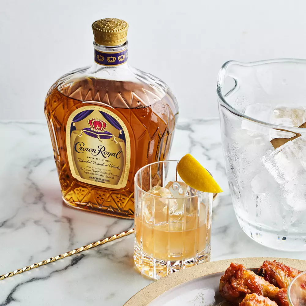 A bottle of Crown Royal whisky, a glass with whisky and ice garnished with a lemon twist, a pitcher of ice, and a plate of food on a marble surface.