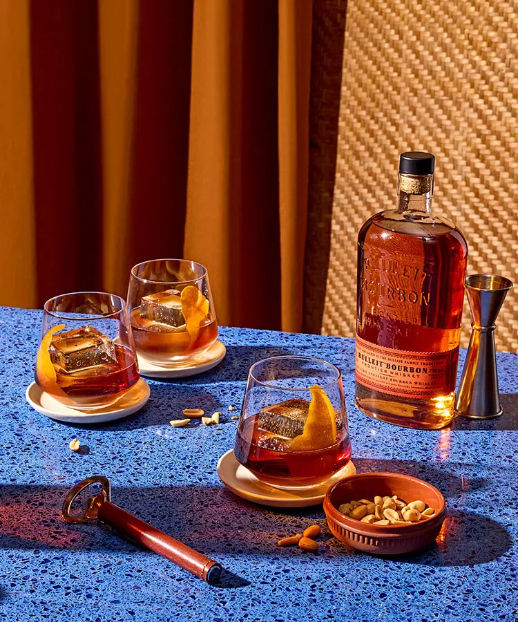 A bottle of Bulleit Bourbon, three bourbon cocktails with orange peels, a jigger, mixed nuts, and a cigar cutter on a blue speckled table.
