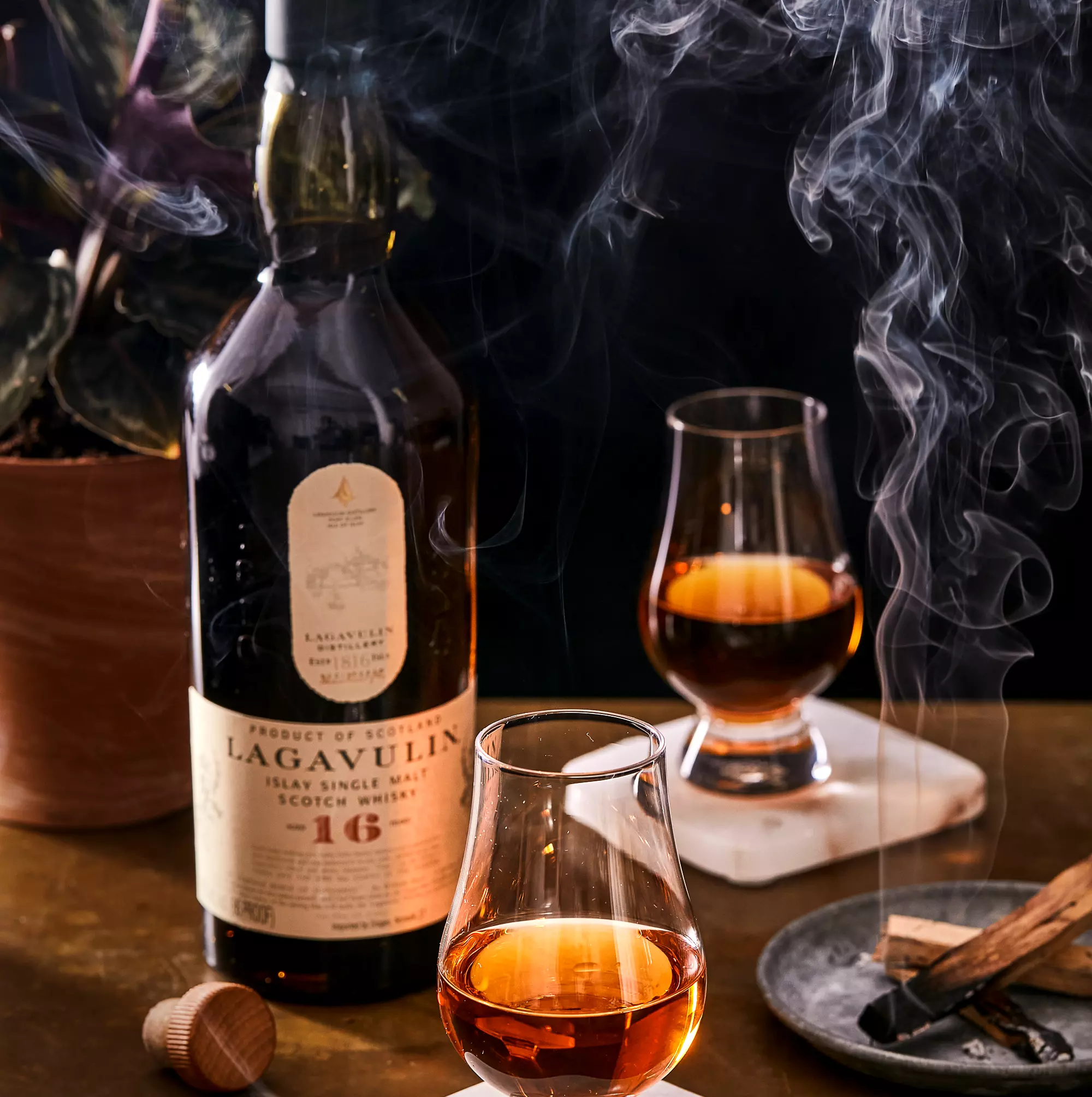 A bottle of Lagavulin 16-year-old Scotch whisky with two filled glasses and swirling smoke on a table, next to a potted plant and a plate with matches.